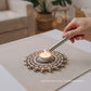 uma base de vela rústica estilo mandala feita em madeira com uma vela ao centro e uma maão feminina a acender a vela