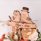 Topo de bolo com pássaros em madeira e nome "Filipa & João", entre flores artificiais coloridas.
