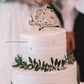 Topo de bolo personalizado com passarinhos de madeira e nome "Filipa & João" sobre bolo de chocolate com açúcar. Fundo decorativo suave.